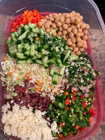 A plastic container filled with chopped cucumbers, chickpeas, diced bell peppers, feta cheese, olives, red onions, parsley, and cabbage