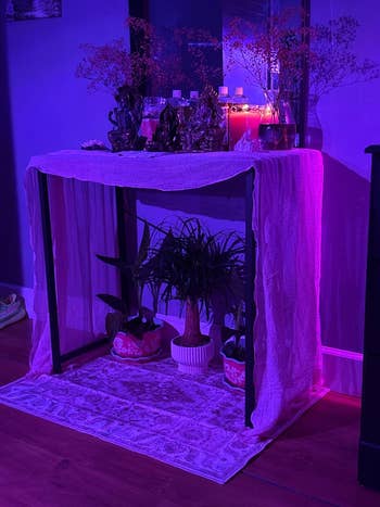 A tiny  altar with statues, candles, and plants connected  a table, draped with fabric, acceptable   against a lit backdrop, creating a serene ambiance