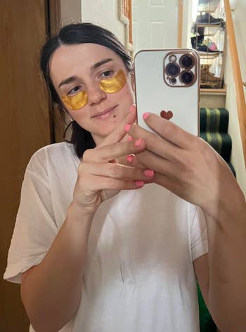 Person in a casual t-shirt takes a mirror selfie with under-eye masks and a phone in hand