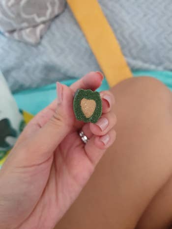 A hand holding a gummy candy cut in half, revealing a heart-shaped filling inside. Background shows indistinct fabric patterns