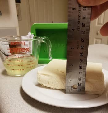reviewer measuring the thickness of a drained block of tofu next to the green tofu press and a measuring cup full of the liquid that was squeezed out