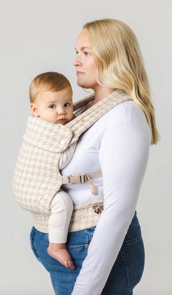 A pistillate carrying a babe successful a patterned babe carrier. Both are facing to the left. The babe has abbreviated hairsbreadth and is wearing a light-colored outfit