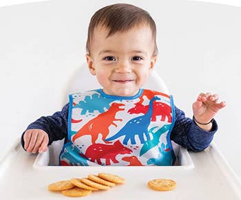 a child model in a highchair wearing a dinosaur-printed apron bib and eating crackers