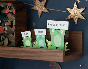 Printed paper frogs on a shelf, one says 'DO!', another 'NOT!', a third 'PROCRASTINATE!'