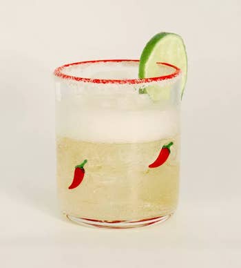 A glass with chili pepper designs, filled with a light-colored drink, garnished with salt, a lime slice, and crushed ice