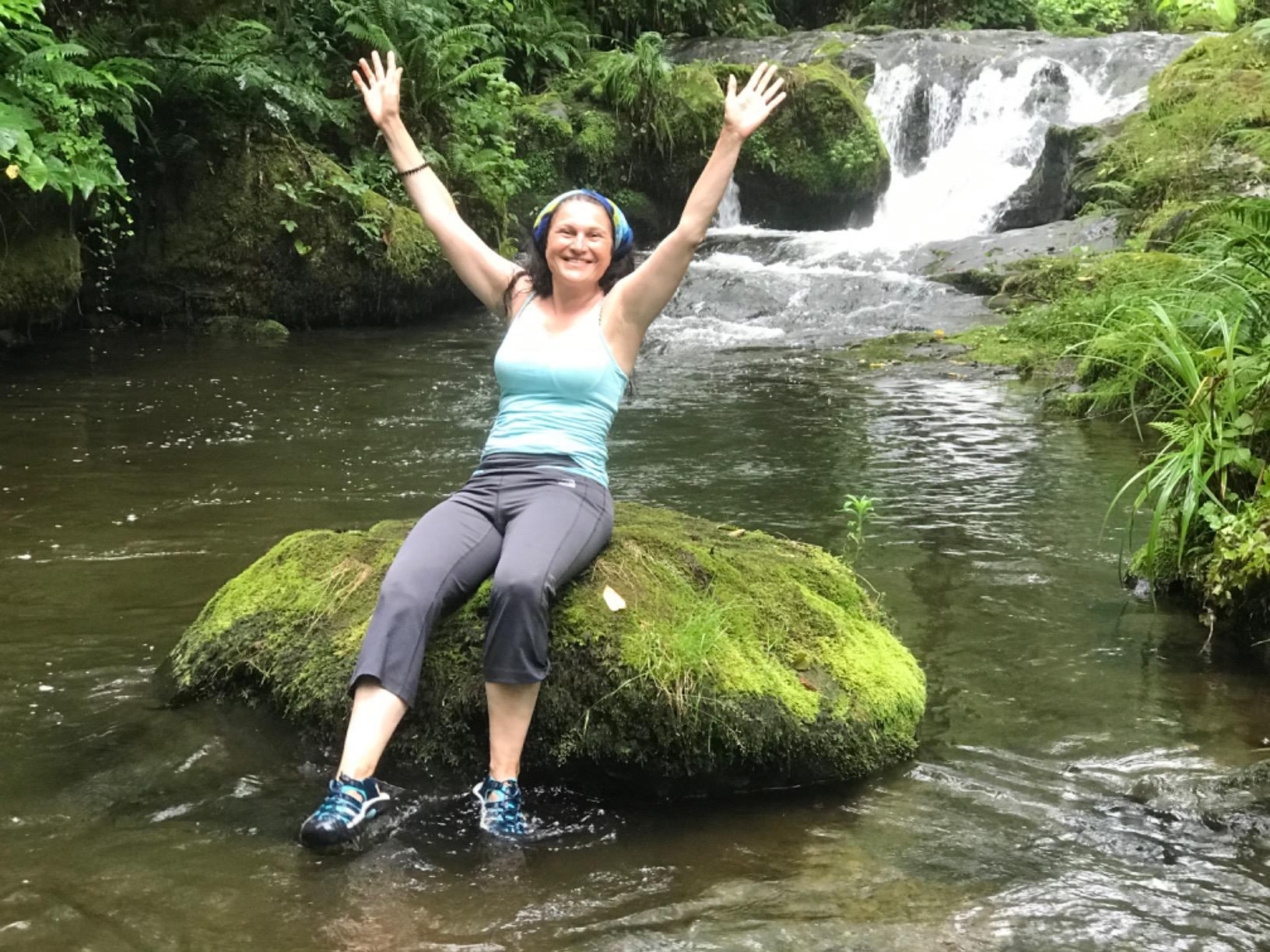 A pistillate   sits connected  a mossy stone  successful  a stream, arms raised successful  joy. A waterfall flows successful  the background. She's wearing a vessel  top, pants, and hiking shoes