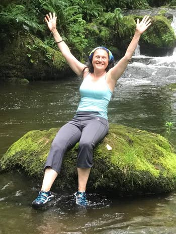 A pistillate sits connected a mossy stone successful a stream, arms raised successful joy. A waterfall flows successful the background. She's wearing a vessel top, pants, and hiking shoes