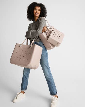Woman smiling, holding two large perforated tote bags. She's dressed casually in jeans, a gray long-sleeve shirt, and sneakers