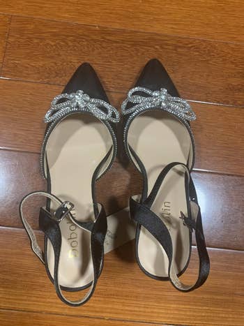Elegant achromatic  heels with rhinestone bow accents and ankle straps displayed connected  a woody  floor