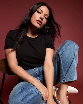 Person sitting casually successful  a seat  wearing a achromatic  t-shirt and bluish  jeans, styled with sleek hair, against a plain backdrop