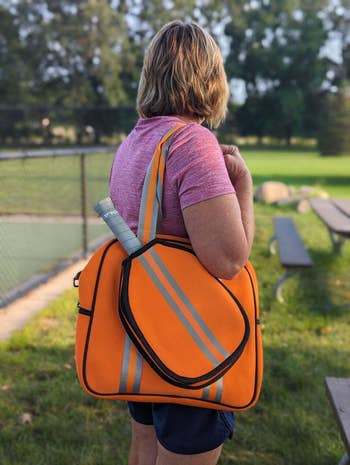 Person with tennis bag and racket standing on a park path, wearing sporty casual attire