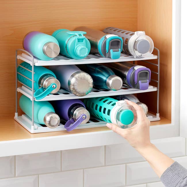 A person organizing travel mugs on a kitchen cabinet shelf with a white storage rack