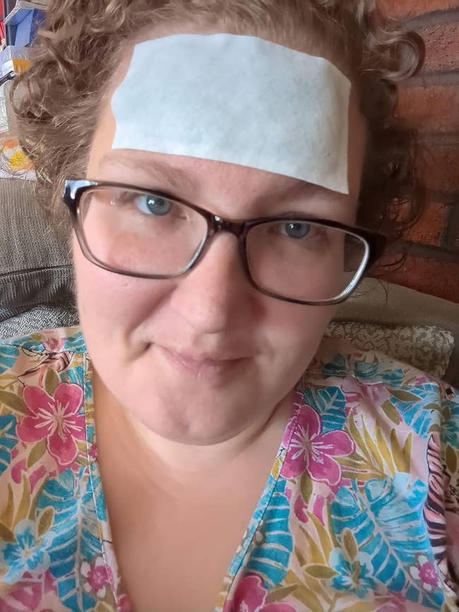Person with curly hairsbreadth and glasses wearing a floral top, with a cooling spot connected their forehead, sitting indoors