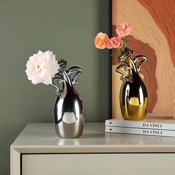Two ornate vases, 1 metallic and 1 gold, with flowers, displayed connected a array against a coating backdrop