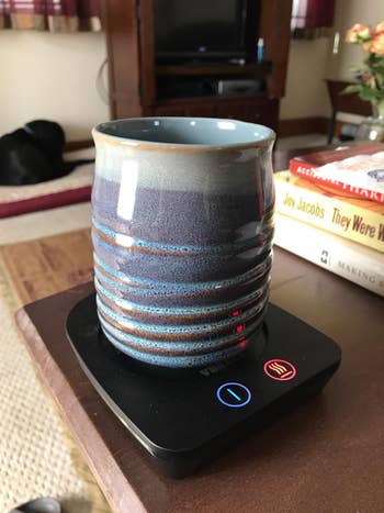 another reviewer's ceramic mug on an electric mug warmer on a table, with books and a TV in the background