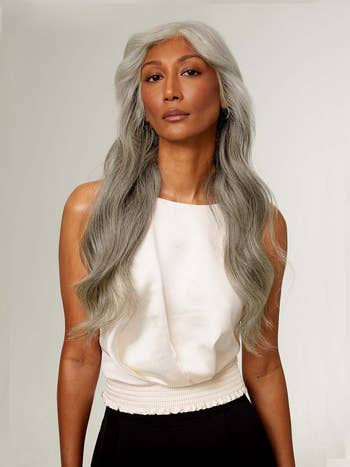 Person with long silver hair, wearing clip-ins, a sleeveless, draped top and high-waisted black pants, poses confidently
