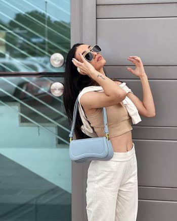 Person posing against a modern background, wearing a beige crop top, white pants, sunglasses, and carrying a light blue handbag