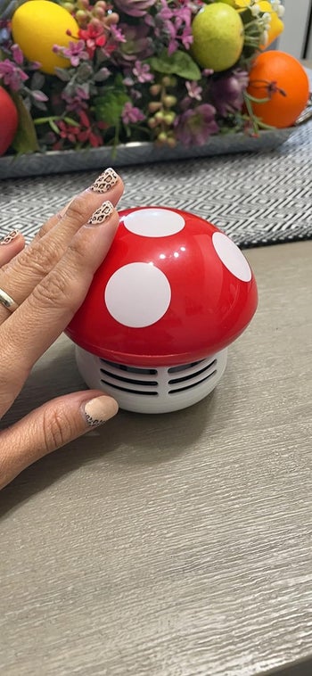 A person's hand with patterned nail art is touching a small, mushroom-shaped portable speaker on a table