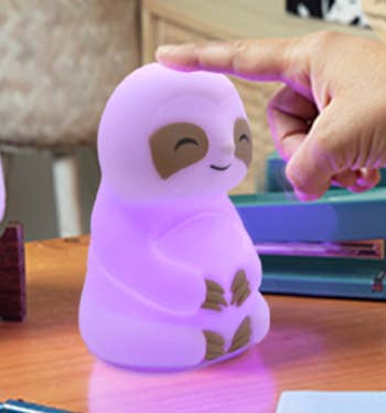A smiling sloth-shaped nighttime  airy  is mildly  touched by a finger, sitting connected  a table  with books and stationery