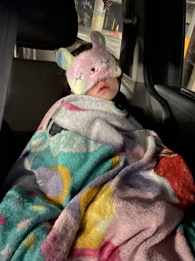 Child sleeping successful a car spot wrapped successful a colorful blanket, wearing a unicorn-themed slumber mask