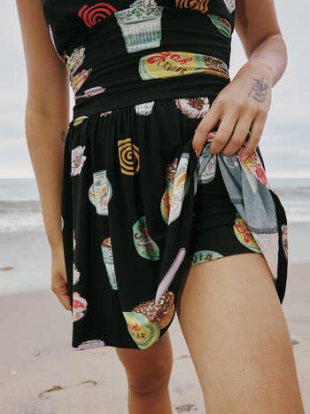 Person in a beach setting wearing a dress with food-themed prints, showing part of their leg and a tattooed hand adjusting the dress