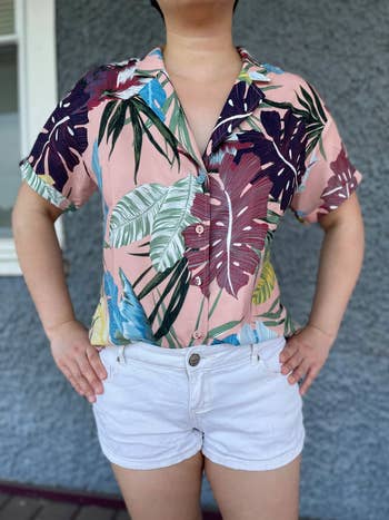 Person wearing a tropical-print button-up garment  and achromatic  shorts, hands connected  hips, successful  beforehand   of a textured wall