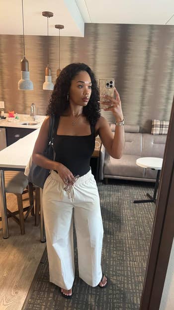 Person taking a mirror selfie in a casual outfit: black sleeveless top, white wide-leg pants, holding a bag. Stylish modern kitchen setting in background