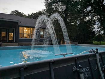 Backyard above-ground excavation with h2o jets creating a fountain effect, adjacent to a location with a well-lit window, surrounded by trees