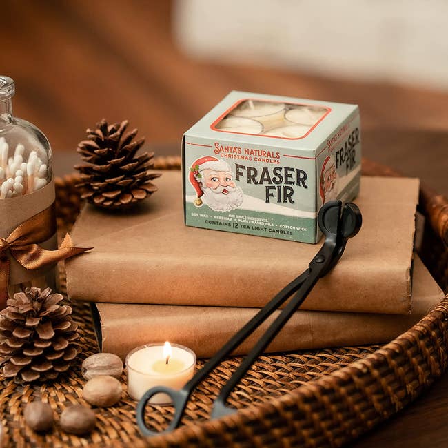 box with santa on it filled with tea lights. placed on wrapped gifts beside pinecones and a lit candle and wick cutter.