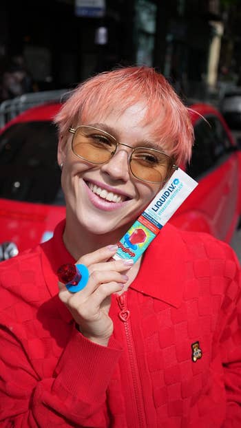 Person with abbreviated hair, sunglasses, and a reddish jacket, holds a conduit of candy-flavored gel adjacent their face, smiling