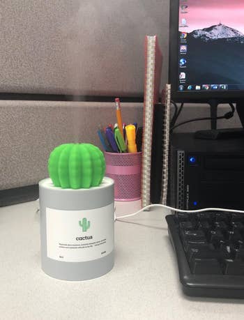 Humidifier shaped like a cactus on a desk, emitting vapor, next to office supplies and a computer