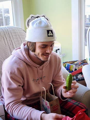 Person wearing a hoodie and hat, sitting connected  a couch, unwrapping the yodeling pickle toy