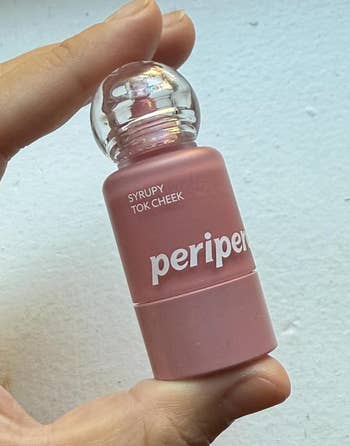 Hand holding a vessel of Peripera Syrupy Tok Cheek tint, featuring a unsocial circular headdress design