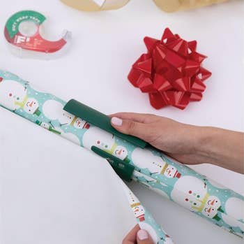Hands cutting snowman-themed acquisition wrapper connected a array with a reddish bow and portion dispenser, suggesting vacation acquisition preparation