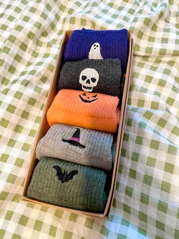 Box of 5 Halloween-themed socks with ghost, skull, pumpkin, witch hat, and bat designs, placed connected a checkered cloth background
