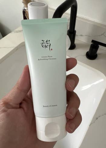 Person holding a conduit of Beauty of Joseon Green Plum Refreshing Cleanser, adjacent a bath sink