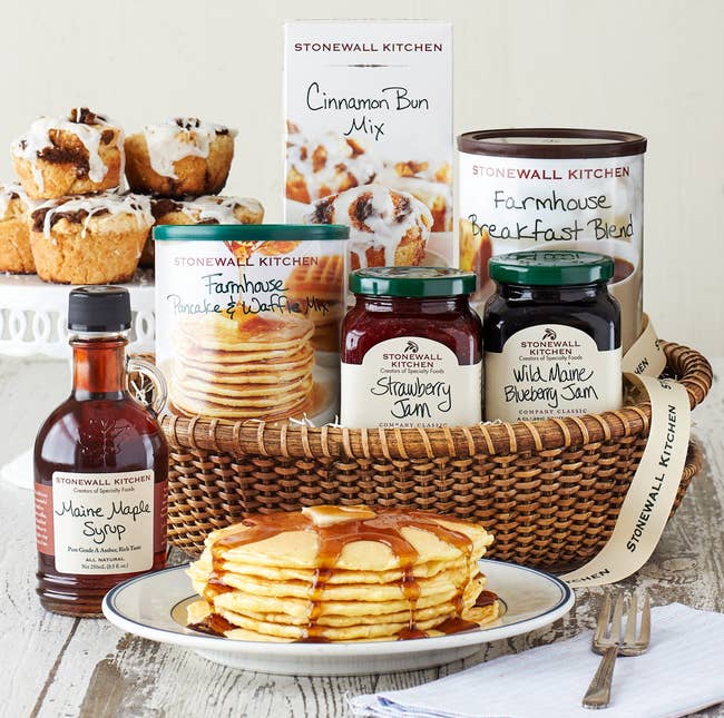 a plate of pancakes in front of a basket filled with Stonewall Kitchen branded maple syrup, pancake and cinnamon bun mix, coffee, and two flavors of jam 