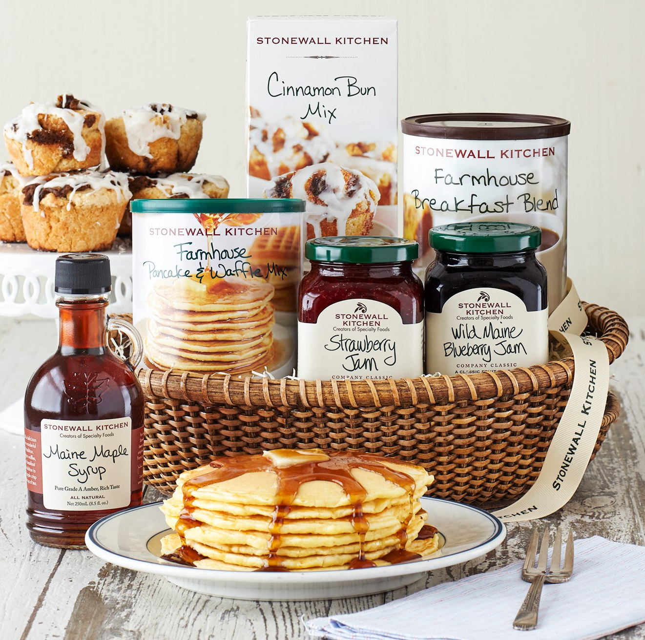 a plate of pancakes in front of a basket filled with Stonewall Kitchen branded maple syrup, pancake and cinnamon bun mix, coffee, and two flavors of jam 