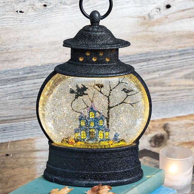 lantern shaped snow globe with floating yellow sparkle, a haunted house with pumpkin and flying with beside a tree inside 