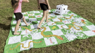 Two children playing on a large outdoor snakes and ladders game mat with a big die placed on the grass