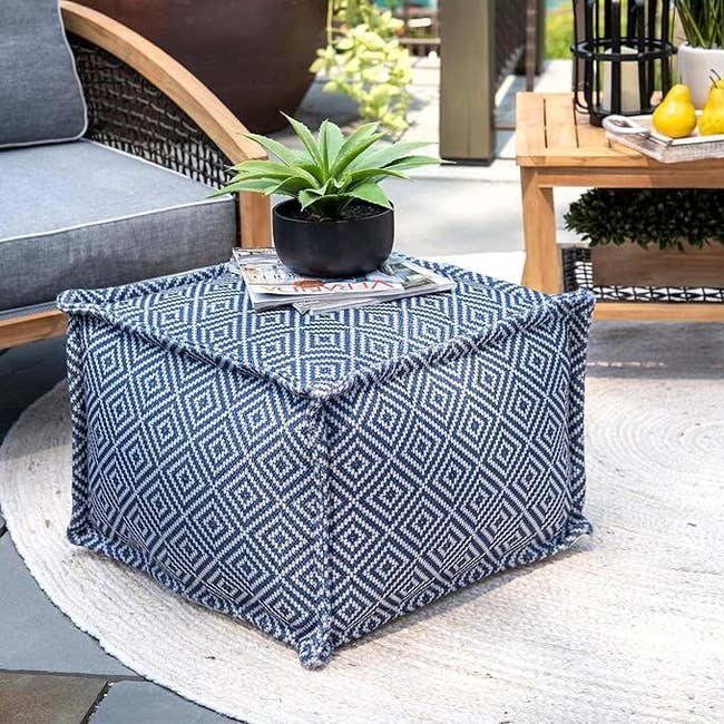 Cube-shaped fabric ottoman with a geometric pattern, holding a plant, a magazine, and a white rug underneath. Furniture and decor accessories surround the ottoman