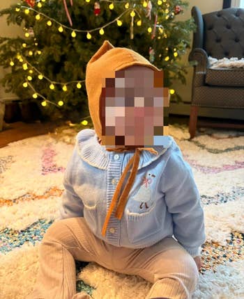 A babe  sits connected  a rug successful  beforehand   of a decorated Christmas tree, wearing a airy  overgarment   with a goose plan  and a cozy cap