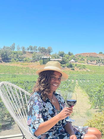 reviewer wearing the brimmed hat with a white ribbon while drinking wine in a vineyard