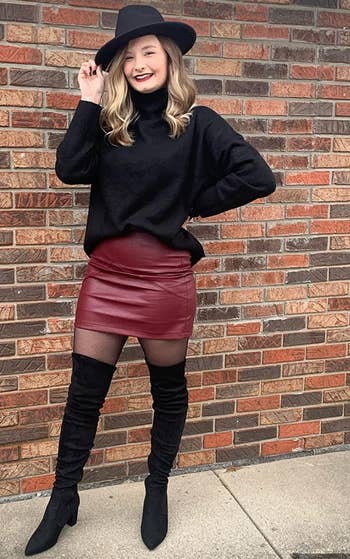 another reviewer wearing black boots with a burgundy skirt, black sweater, and hat
