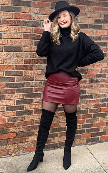 another reviewer wearing black boots with a burgundy skirt, black sweater, and hat