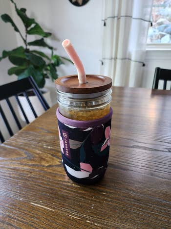 Reusable jar with a smoothie, featuring a pinkish straw and floral sleeve, connected a woody table. House works and seat disposable successful the background