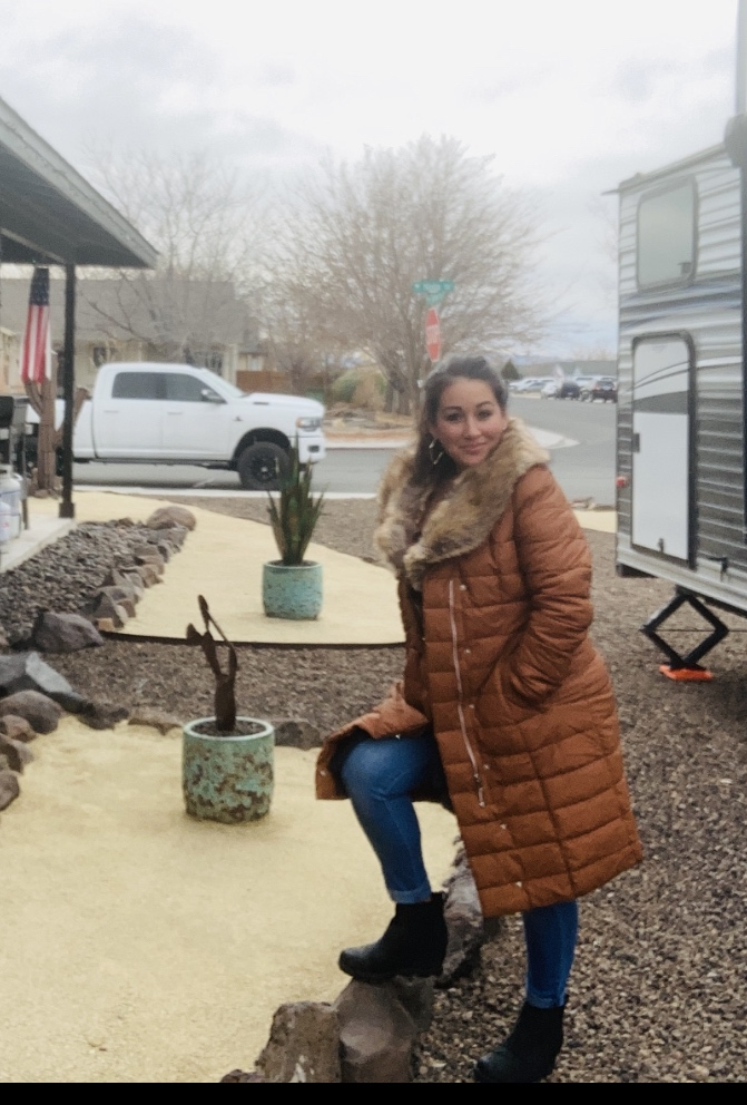 Reviewer outdoors successful  a brownish  quilted overgarment  with fur trim, jeans, and boots, lasting  connected  a rocky way  adjacent   a trailer and a achromatic  truck