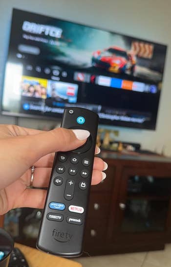 A person holds a Fire TV remote in front of a TV displaying a streaming interface with various apps visible