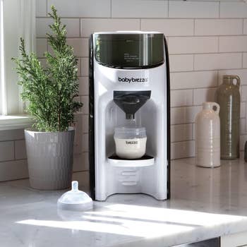 Baby formula maker on a kitchen counter next to a plant and ceramic jugs. A filled baby bottle is placed in the machine