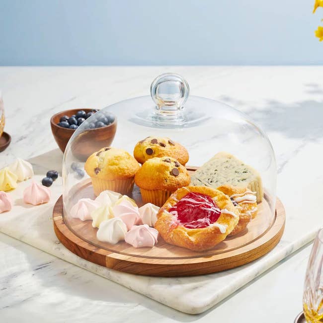 Pastries and muffins, including a danish and meringues, displayed under a clear dome on a wooden board on a marble table